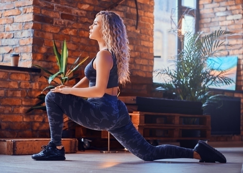 woman is doing aerobic exercise at modern loft. She is wearing sport leggings bra and sneakers. Woman has blond curly hair.
